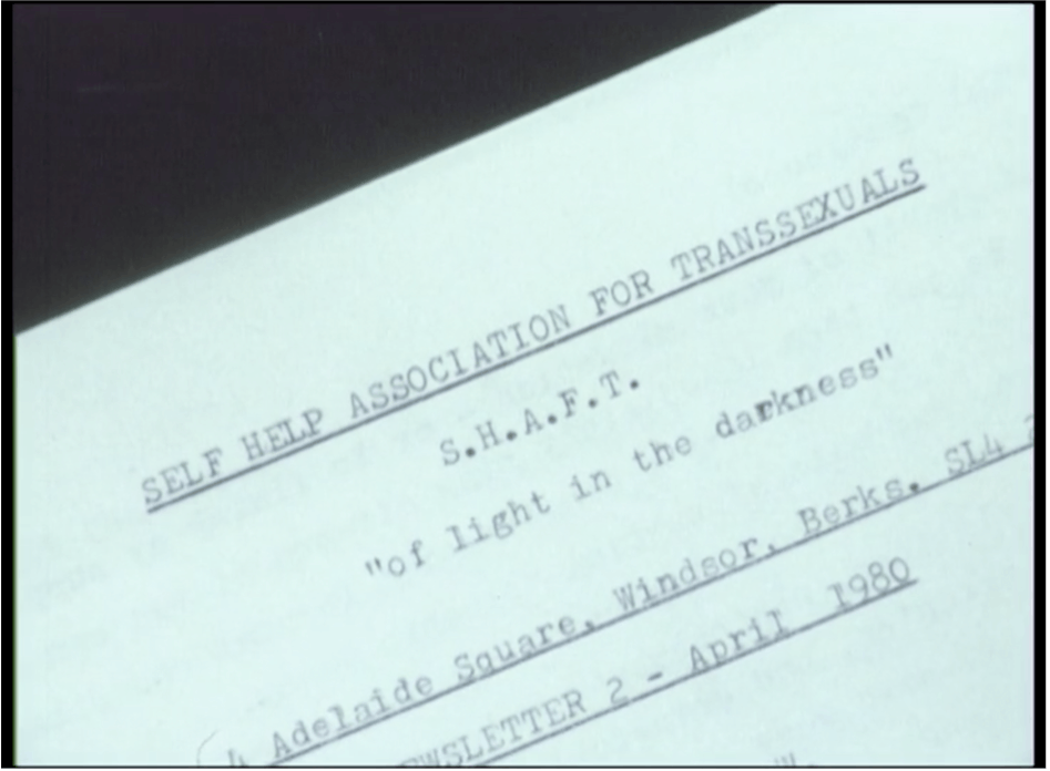 Film still featuring a close-up of a paper document with typewritten text: SELF HELP ASSOCIATION FOR TRANSSEXUALS/ S. H. A. F. T./ "of light in the darkness"/ Adelaide Square, Windsor, Berks, SL4/ Newsletter 2 - April 1980
