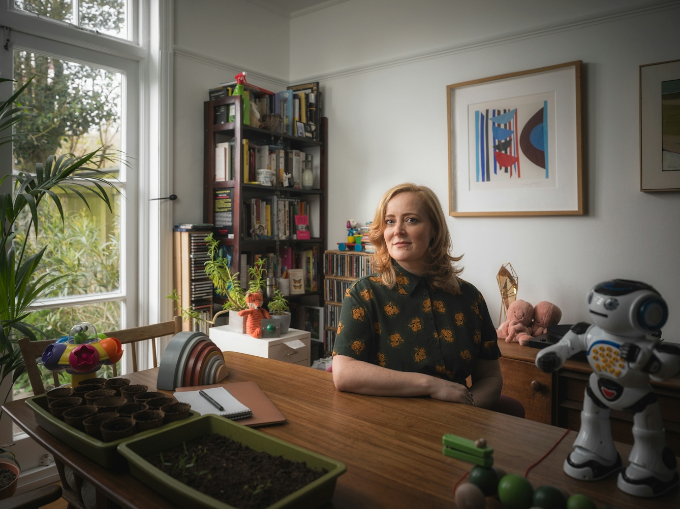 Photographic portrait of a woman in a white dining room, with her arm perched on the table.  Surrounding her are various child’s toys including a robot, a small wooden crocodile and a pink fluffy octopus. There are picture frames on the wall in the background to the right of the frame, a set of shelves with CDs, and some house plants to the left of the frame.  In the foreground to the left, there is a tray of seedlings.