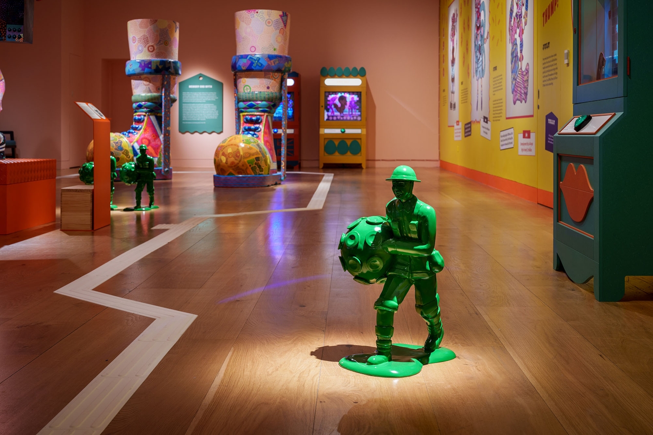 Photo of the exhibition ‘Jason and the Adventure of 254’ showing a sculpture of a “toy soldier” holding a model virus. Behind the sculpture are more artworks and sculptures in the exhibition gallery.