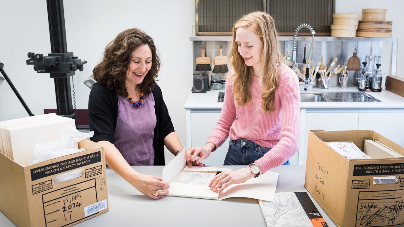 Photograph showing two female conservators standing at a workbench in a museum conservation studio. They are both in the process of carefully opening an archive folder containing a sketch book. To the left and right of them on the workbench are two open cardboard boxes also containing archive material. Behind them part of a photographic copy stand can be seen and a sink area covered in tools and brushes.