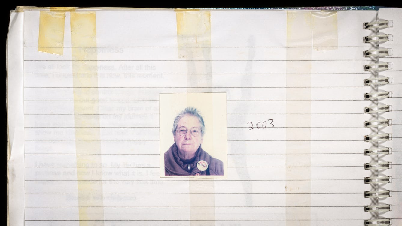 Photograph of the top left hand corner of a spiral bound, lined scrapbook. In the centre of the image is a passport photograph of a woman with greying hair, wearing glasses. Pined to her coat collar is a circular badge with the word 'FORCE' written across it. The photo is held to the page with a long strip of yellowing sellotape. There are several other strips of sellotape attached to the page, all running vertically. Handwritten to the right of the photo is the year '2003'.