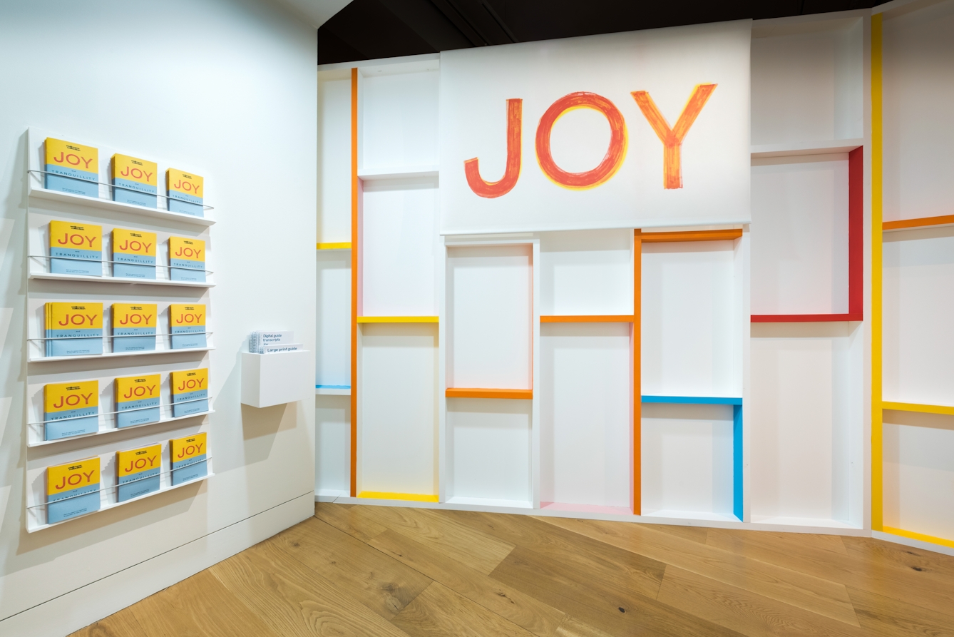 Photograph of an exhibition gallery space showing white walls and a wooden floor. On the right hand wall are 5 narrow shelves holing 15 stacks of an exhibition guide with the word, 'Joy' written in large red letters above a blue band. On the centre and right walls is the large title 'Joy' also written in red and yellow combined colours. Around the title are are 3D grid structure like wall studding spread across the walls. The front edge of this studding is painted in orange, red, blue and yellow colours.