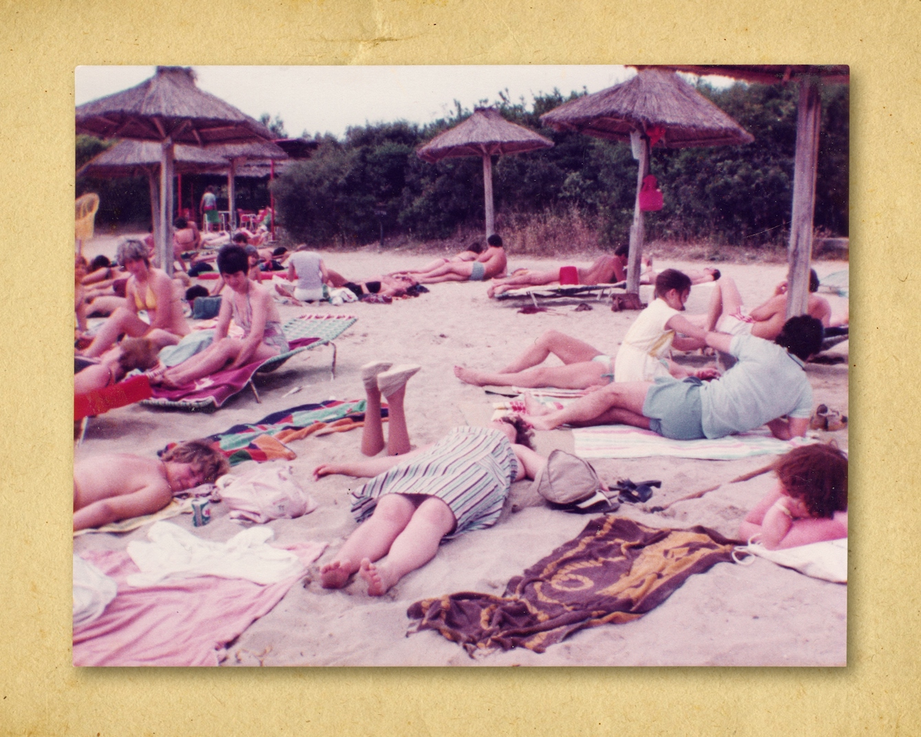 Photograph of a colour photographic print, resting on a brown paper textured background. The print shows a beach scene with many people lying on towels and sun beds enjoying the sun. In the background are parasols made of wooden poles and dried grass umbrellas. In the middle of the scene are a pair of prosthetic legs sticking out from the sand, upside down, feet in the air, disappearing into the sand at the knees.