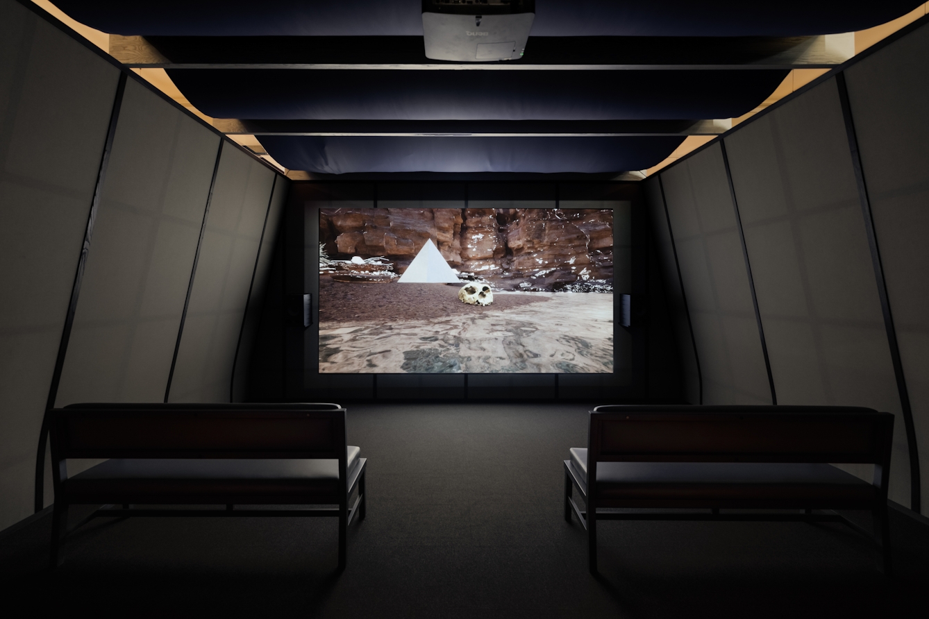 Photograph of a dark exhibition space featuring a projected video on a large screen. Two bench seats are arranged facing the screen. The walls of the room slope inwards and are criss-cross of black wooden battens on grey fabric. The ceiling is a cloth draped over several cross joists. The video shows a computer generated image of a shoreline set against a red rock face. On the sandy beach is a human skull in front of a floating white pyramid shape.