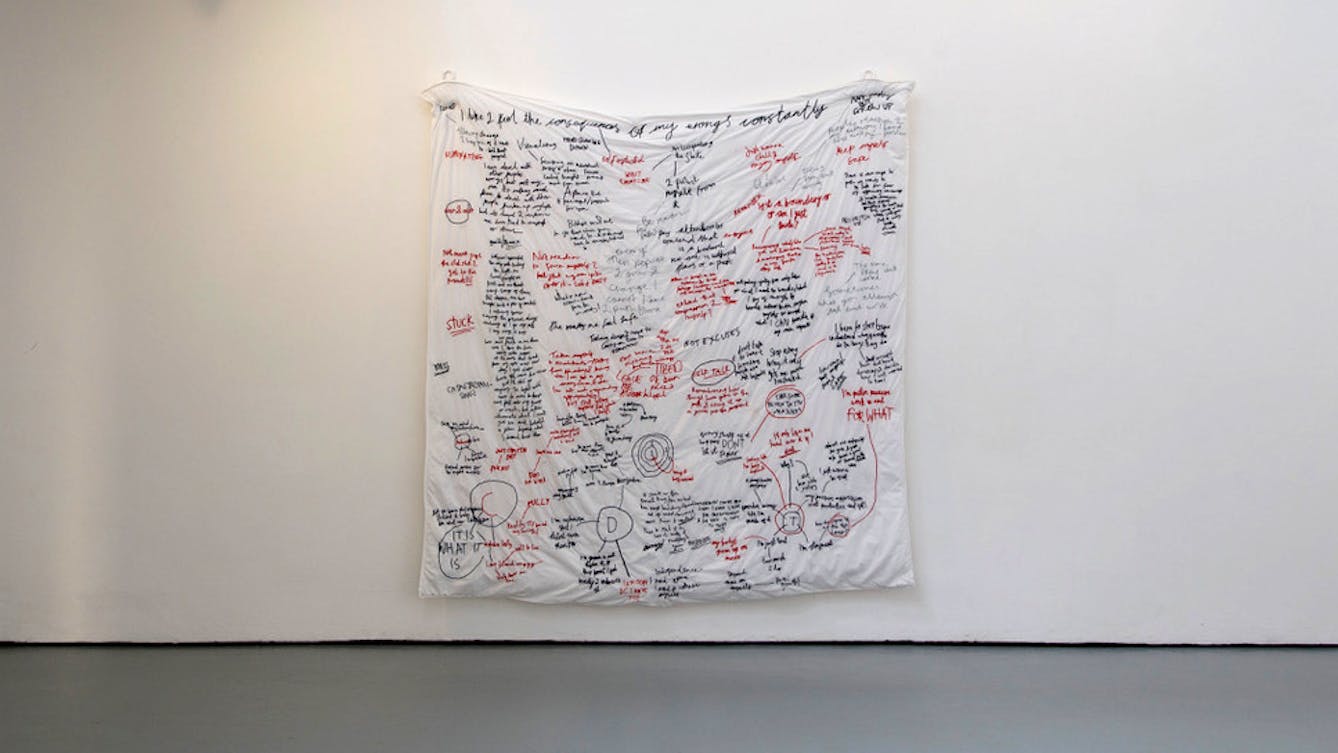 A white duvet cover, embroidered with text in red and black thread hangs on a white gallery wall. 
