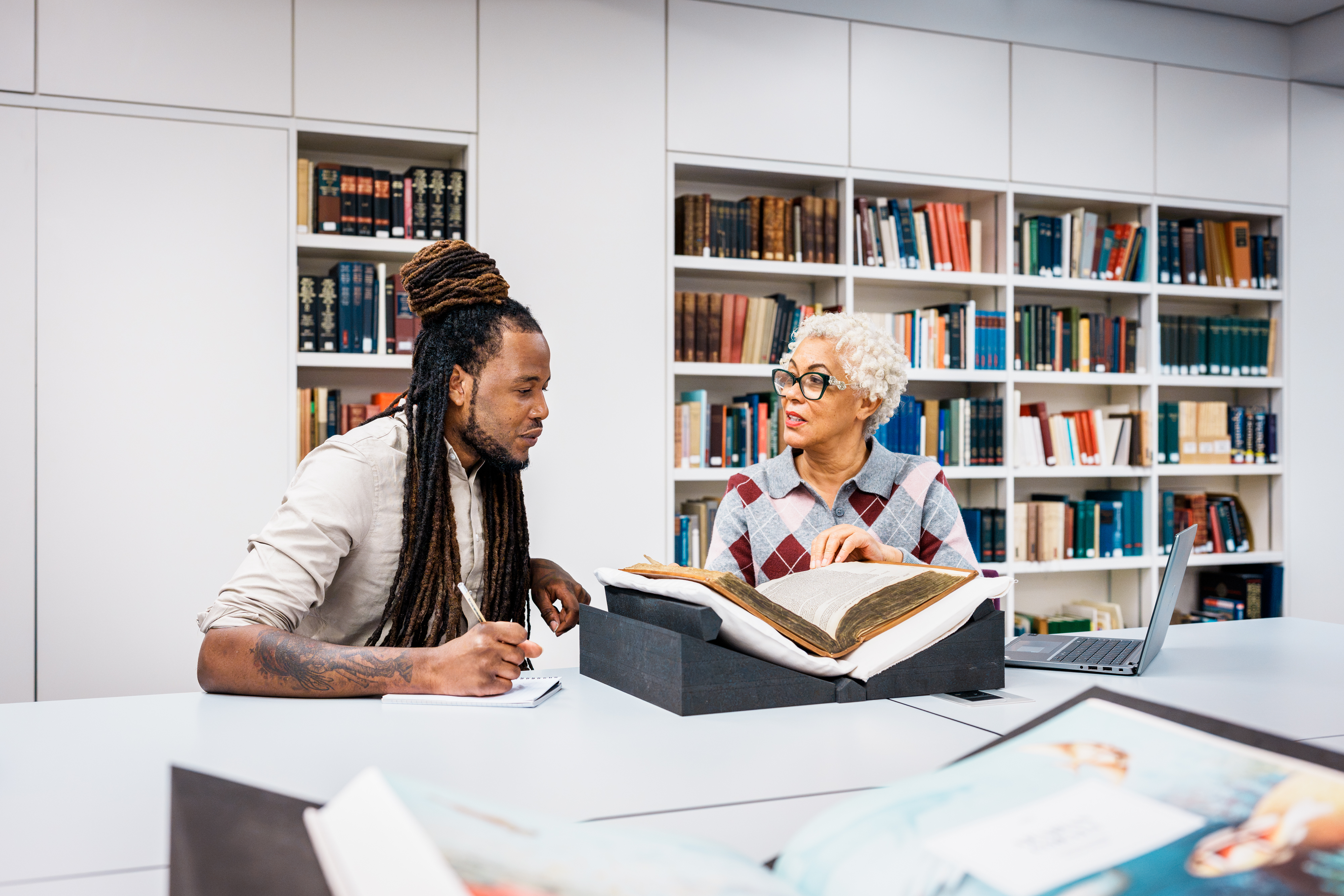 Photograph of 2 library users sat at a table discussing a large fragile bound volume placed on a book rest in front of them.