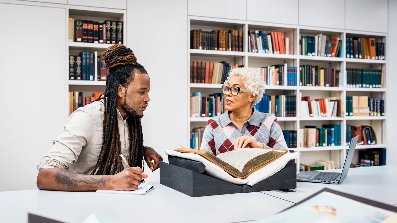 Photograph of 2 library users sat at a table discussing a large fragile bound volume placed on a book rest in front of them.