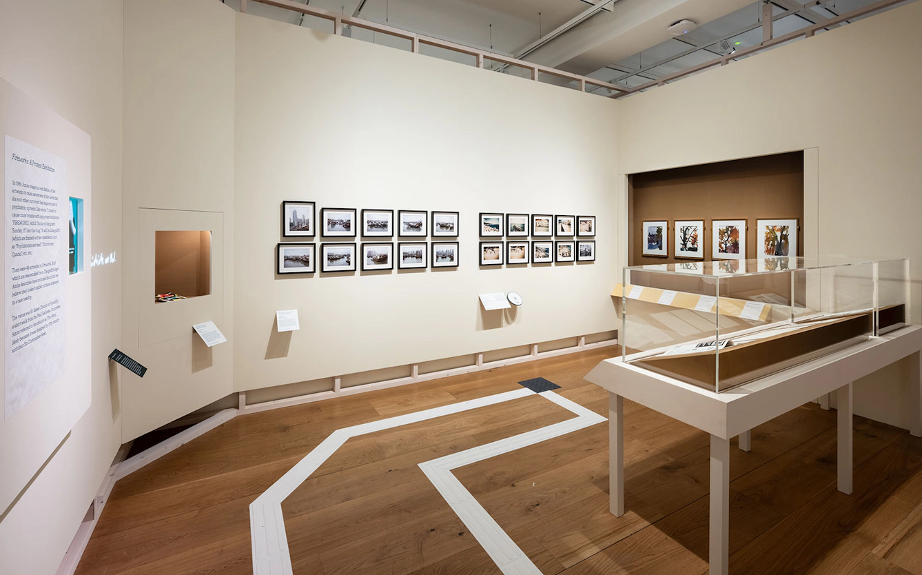 A section of the exhibiton 'Audrey Amiss: the surviving exhibitions' containing a glass enclosed display cabinet and paintings displayed in rows on two walls, and a large text panel on the third wall. The section is called 'Fireworks: A Protest Exhibition'.