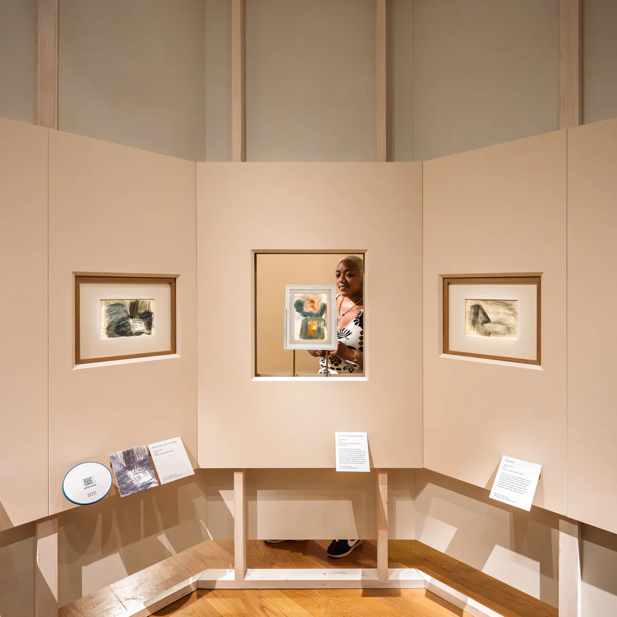 Photograph of a V shaped gallery wall showing 5 painted artworks in frames. The frame in the centre is displayed in a glass window and a visitor can be seen on the other side of he wall.