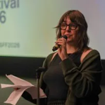 Black and white photo of a woman with glasses and shoulder-length hair standing and giving a talk. She has a microphone in one hand and a sheaf of papers in the other.
