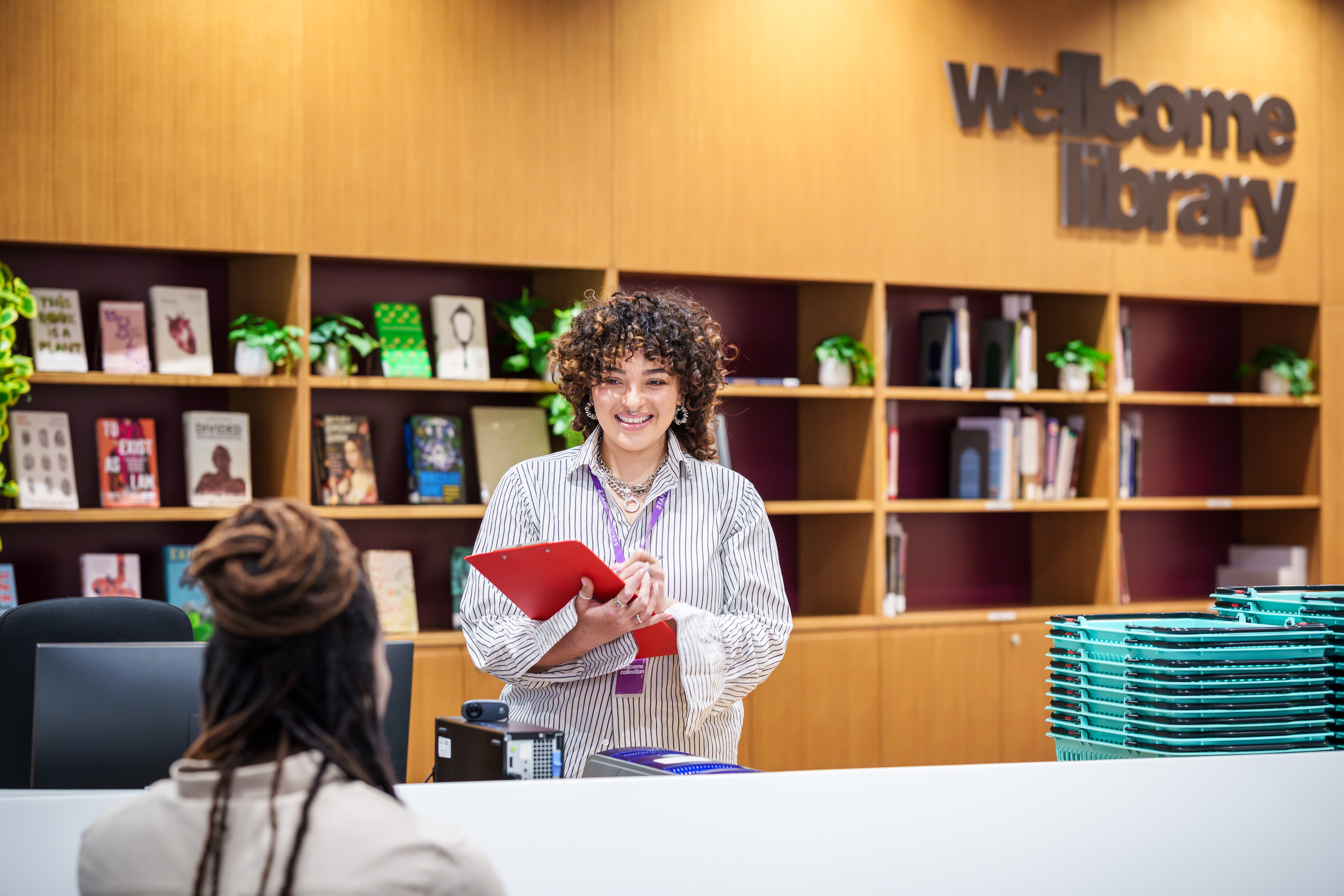 Photograph of a library staff member with a clipboard talking to a library user over the enquiries desk.