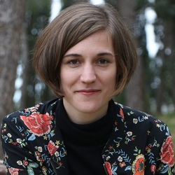 Headshot of a person with short brown hair wearing a black floral jacket, photographed outdoors against a blurred woodland background.