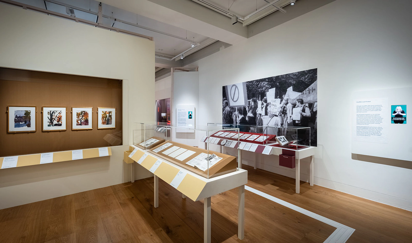 A section of the exhibition 'Audrey Amiss: the surviving exhibitions' containing two glass enclosed display cases and arkworks and photographs displayed on the walls. the section is called 'Graffiti and Protest'