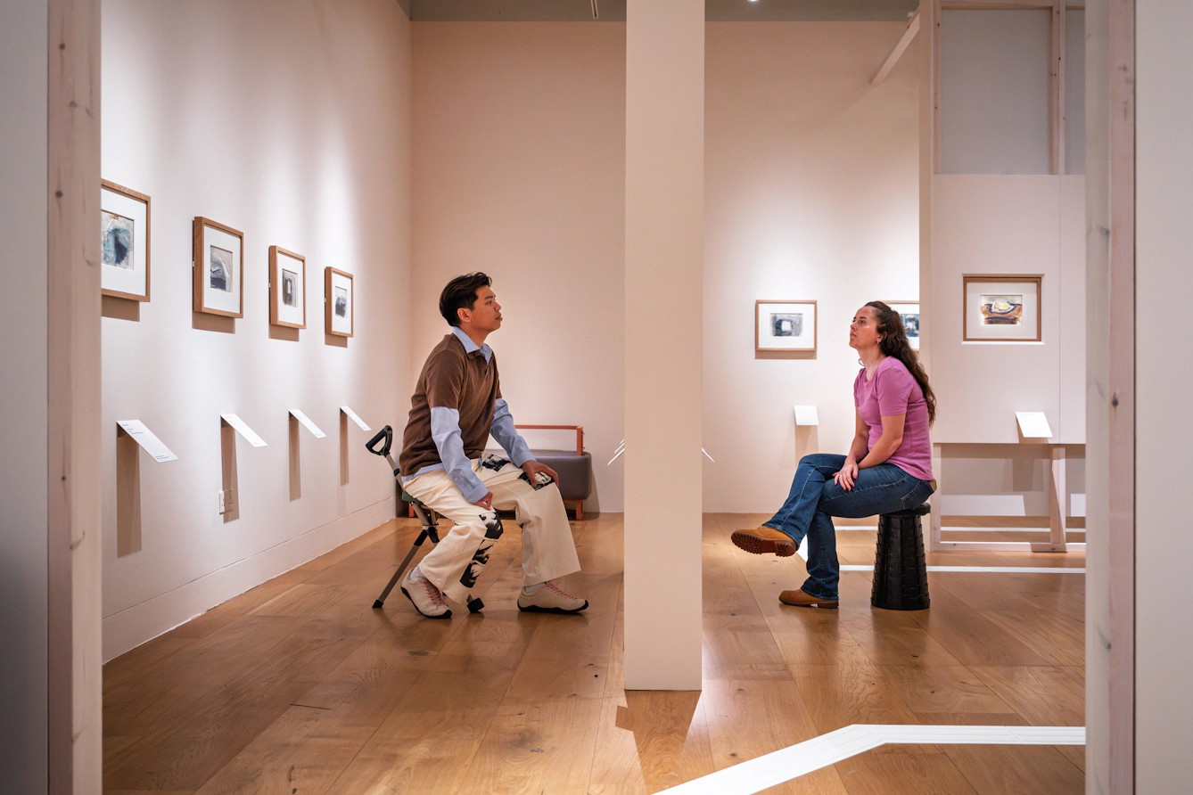 Photograph of two gallery visitors sitting on fold-up chairs either side of a gallery wall.