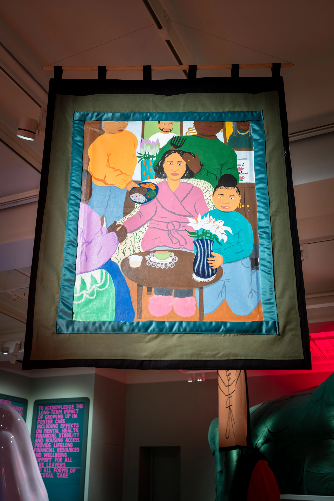 Photograph of a hanging textile artwork showing several people gathered around a seated figure, sharing food, flowers and gestures of care.