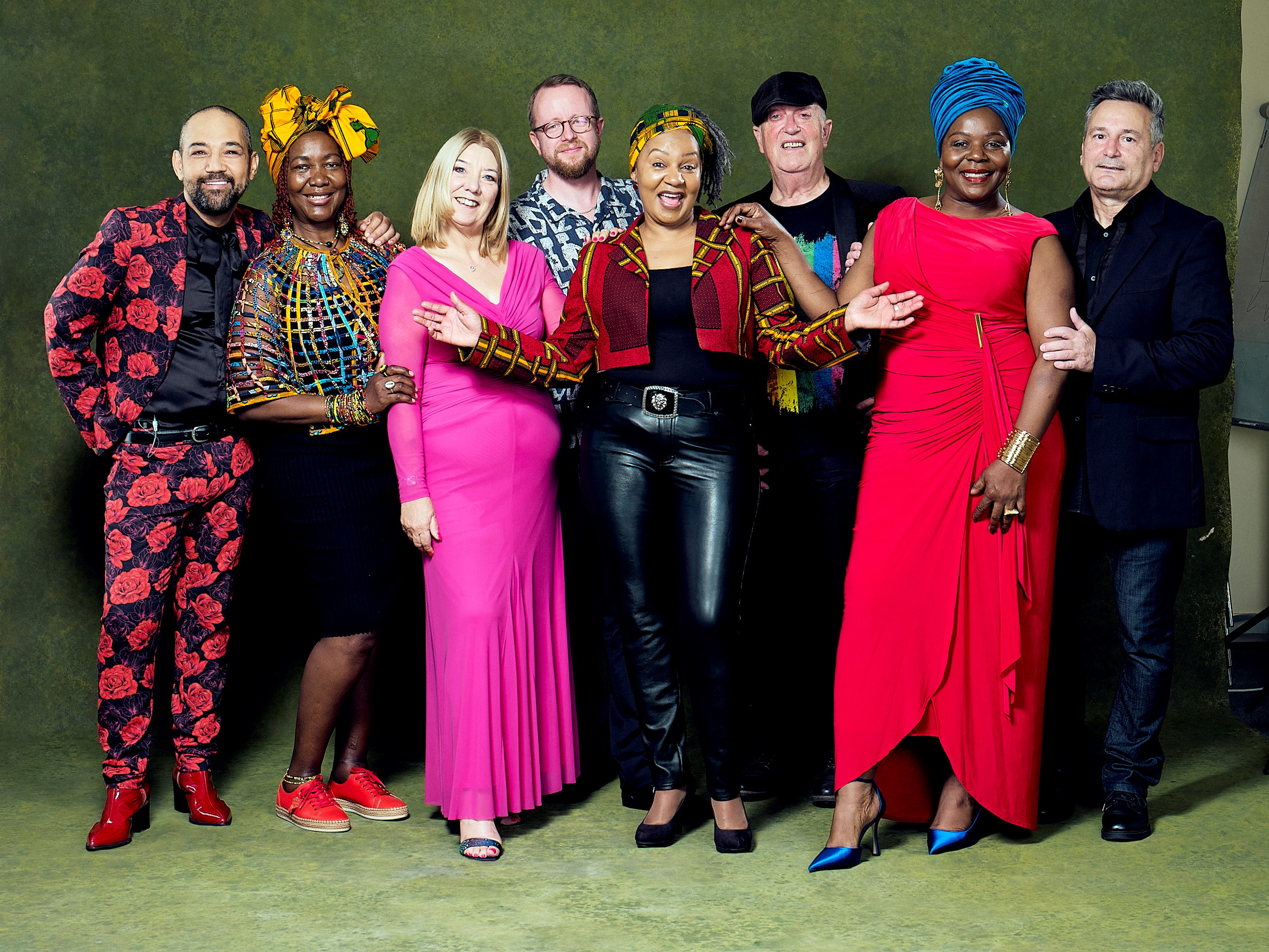 Group portrait of seven people standing together against a green backdrop, wearing colourful patterned and formal clothing.