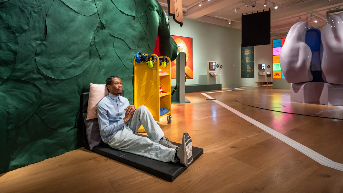 Visitor seated on a floor cushion beside a large sculptural installation, with headphones and sensory resources available nearby.