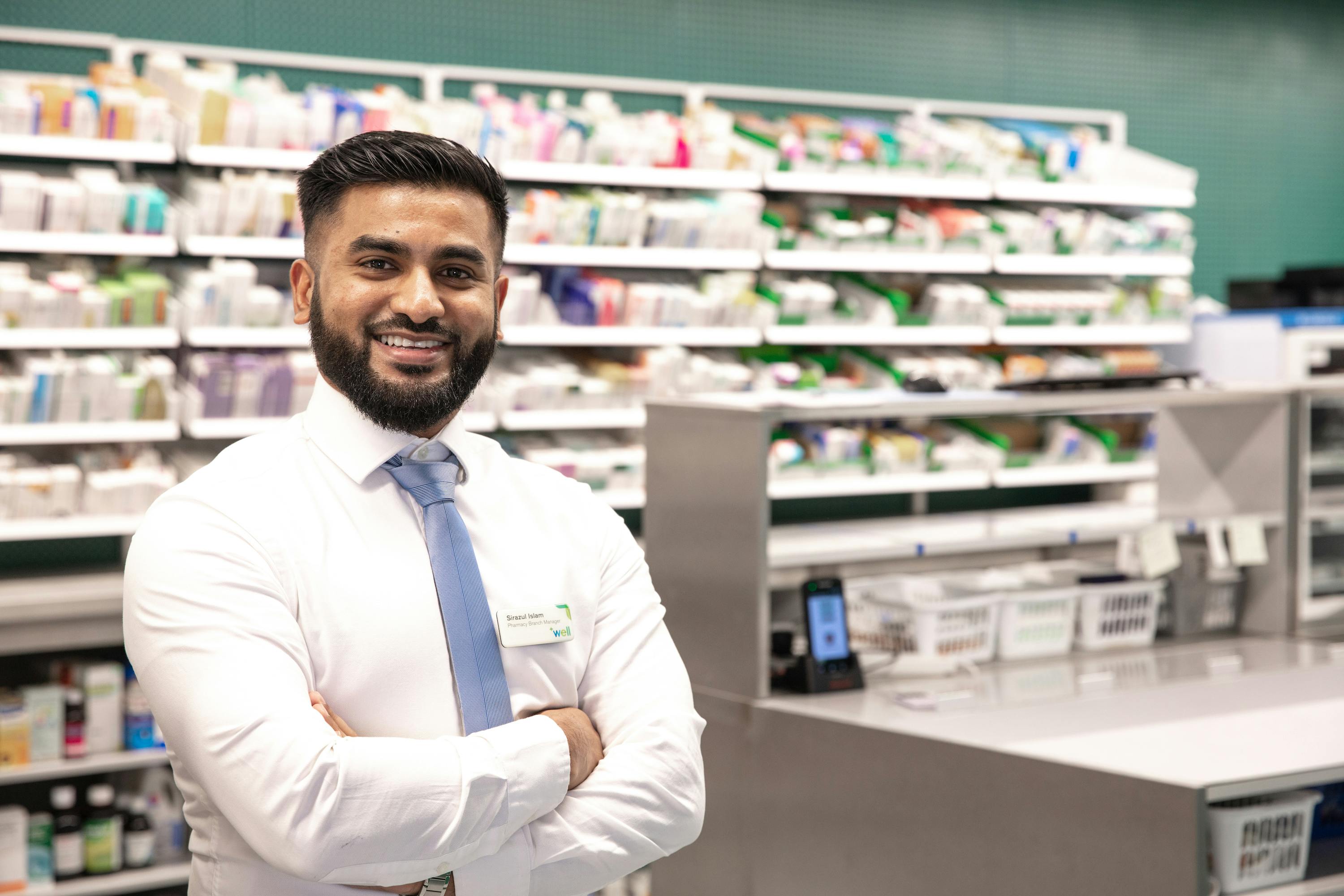 A Well pharmacist standing in front of shelves of medication.