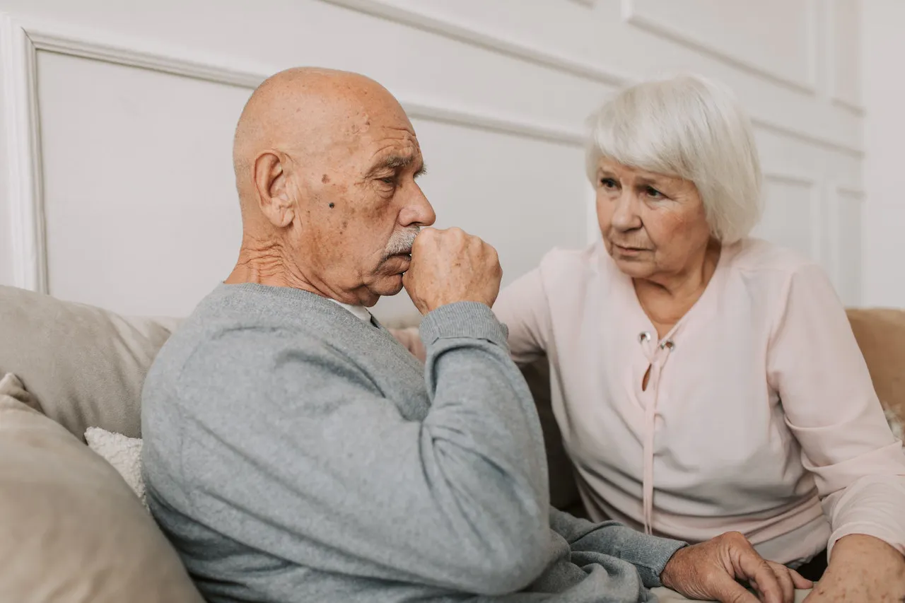 Älterer hustender Mann und ältere Frau sitzen nebeneinander auf dem Sofa