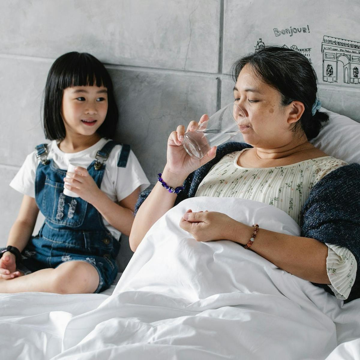 Mutter und Tochter sitzen nebeneinander im Bett während die Mutter mithilfe eines Glas Wassers Medikamente schluckt
