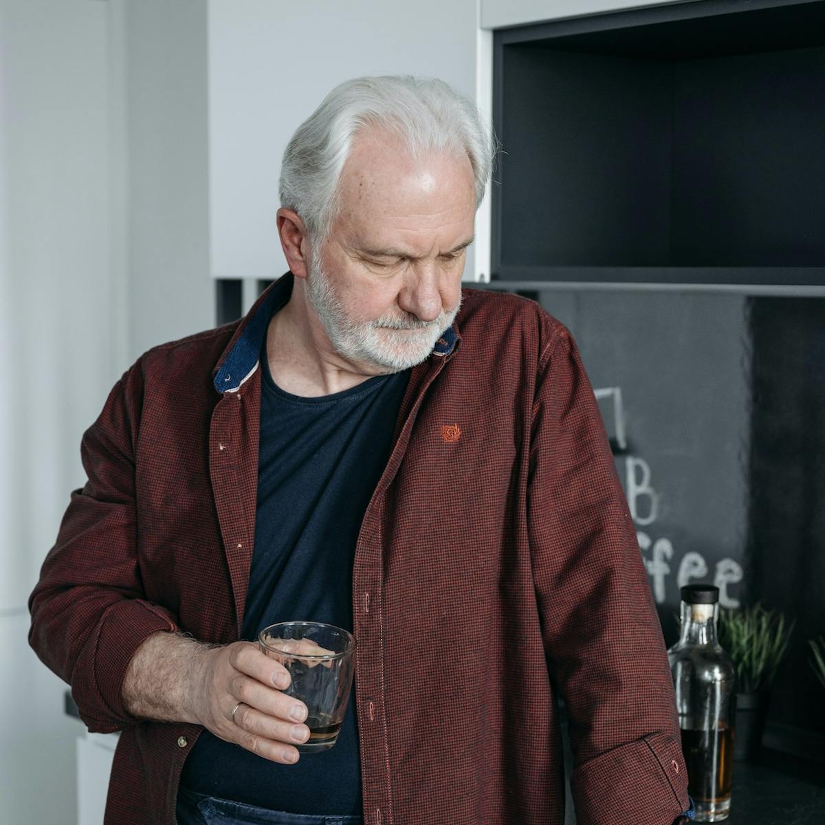 Älterer Mann steht in der Küche mit einem Glas Wasser in der Hand und schaut traurig nach unten