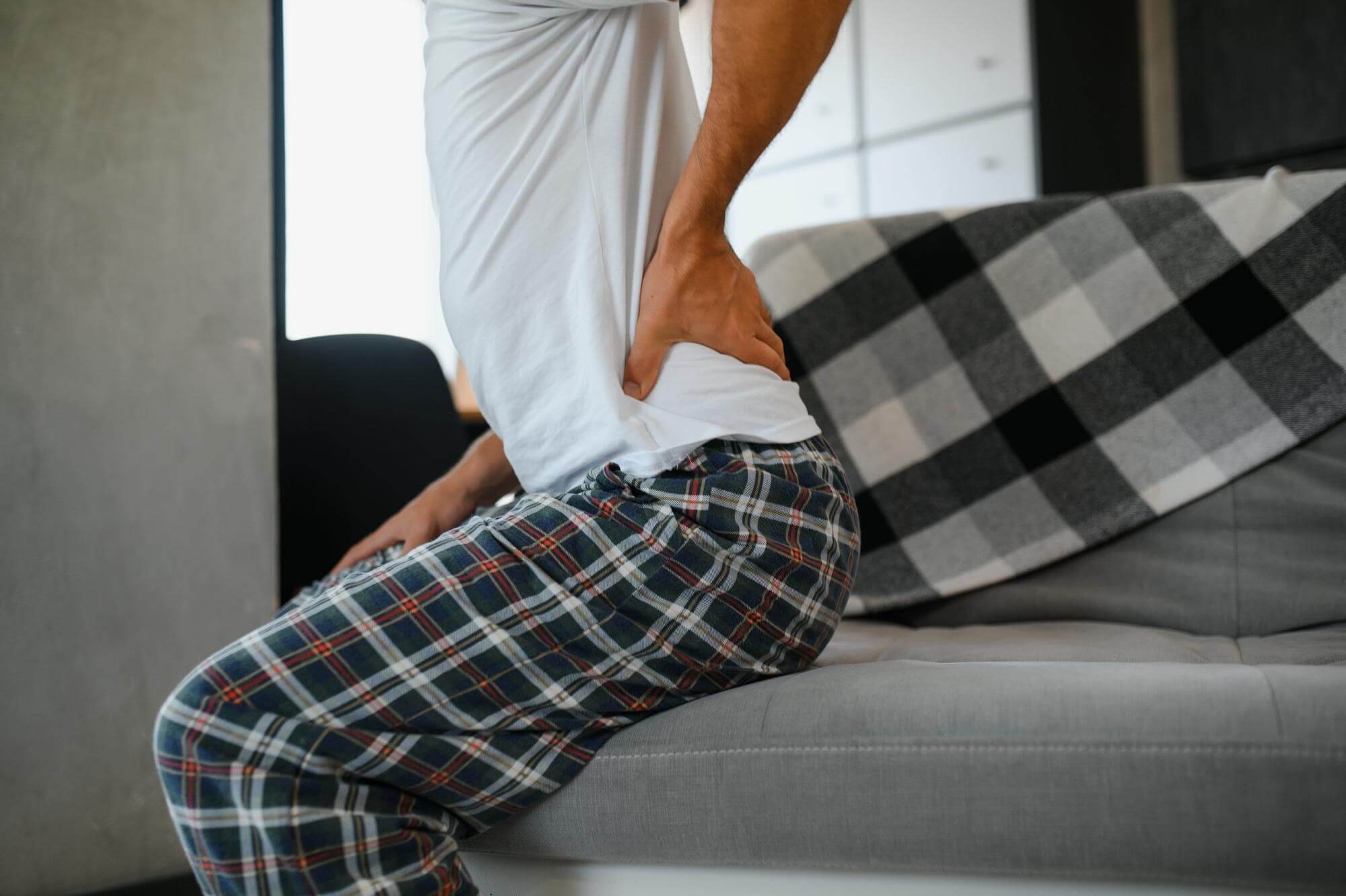 Junger Mann sitzt auf dem Sofa und fasst sich an den unteren Rücken