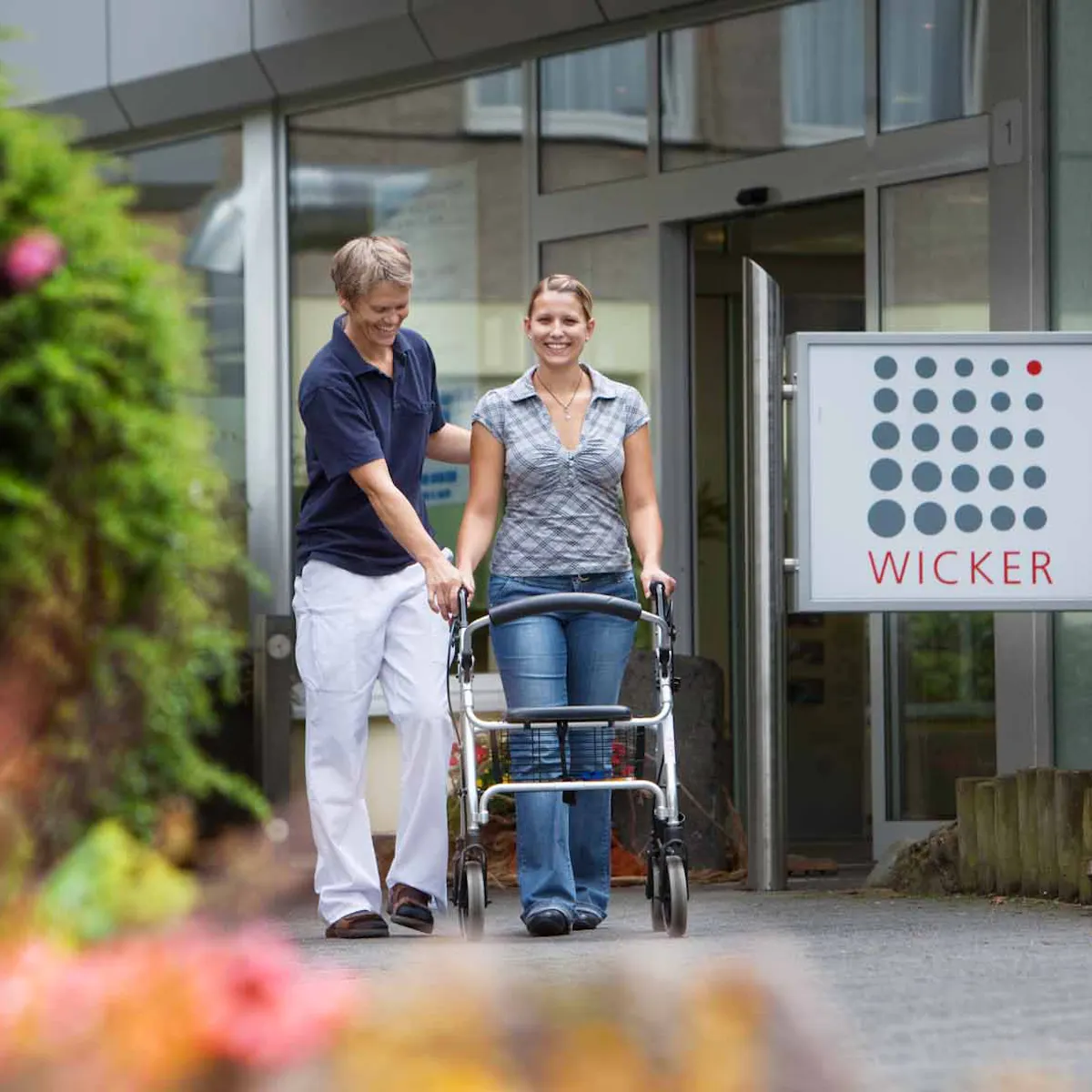 Therapeutin unterstützt Patientin mit Rollator vor dem Eingang der Hardtwaldklinik I in Bad Zwesten