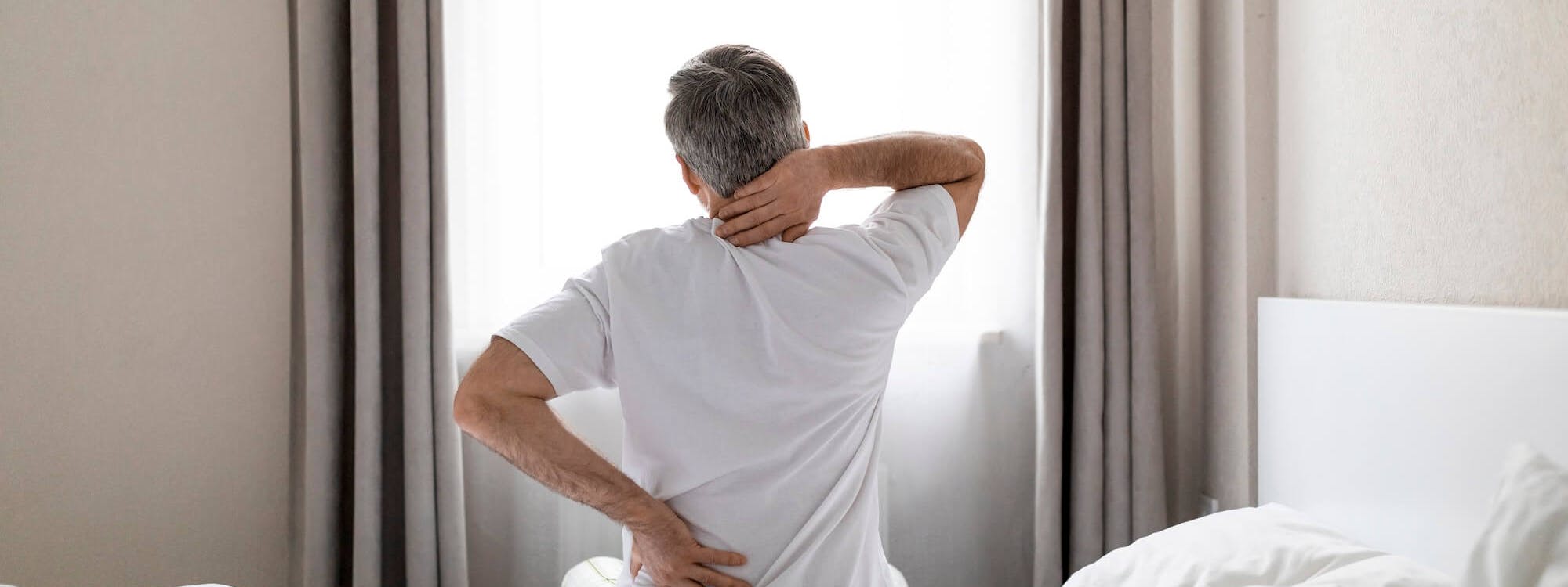 Mann sitzt auf Bettkante und fasst sich an den Rücken Mann sitzt auf Bettkante und fasst sich an den Rücken