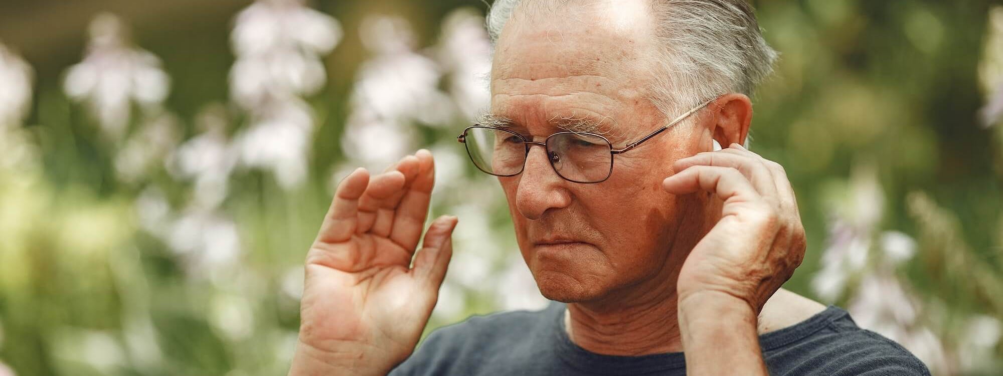 Älterer Mann sitzt in Parkumgebung und hält die Hände an die Ohren