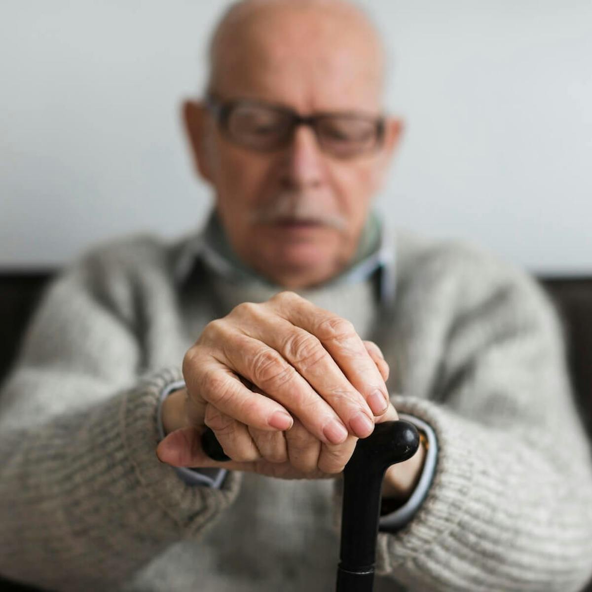 Älterer Mann sitzt auf einem Sofa und stützt seine gefalteten Hände auf einen Gehstock