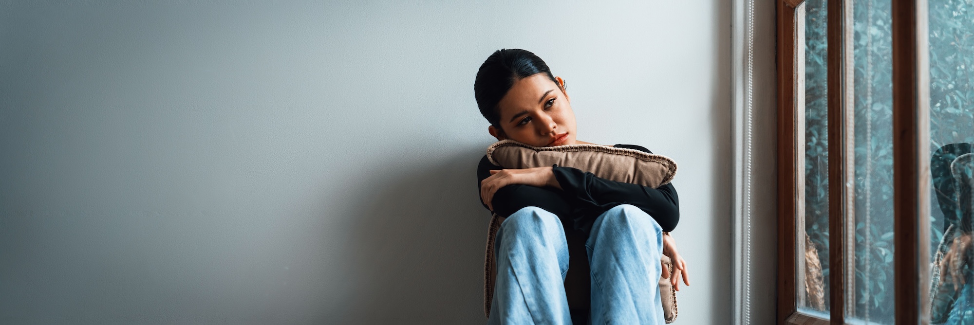 Frau sitzt traurig am Fenster und legt Kopf und Arme auf die angewinkelten Knie