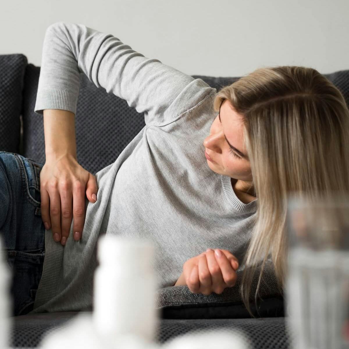 Frau liegt auf dem Sofa und fasst sich mit schmerzverzerrtem Gesicht mit der Hand an den Bauch