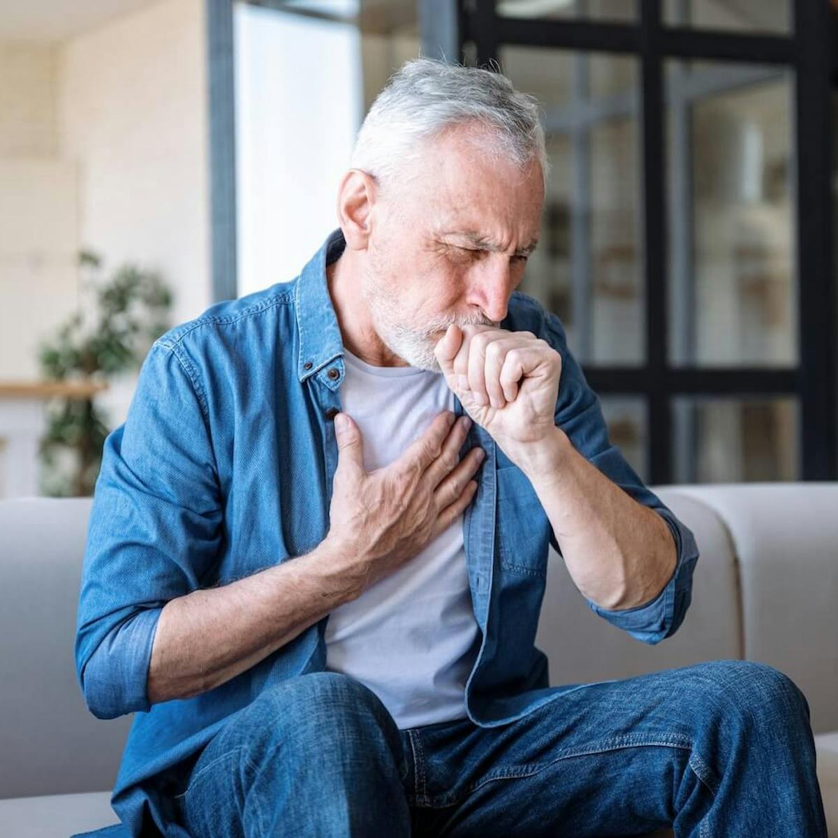 Mann sitzt auf dem Sofa und hustet und hält seine Hand auf die Brust