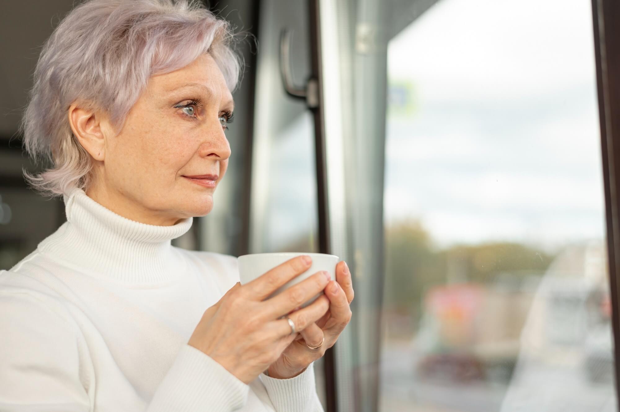 Ältere Frau steht mit einer Tasse in der Hand am Fenster und schaut heraus