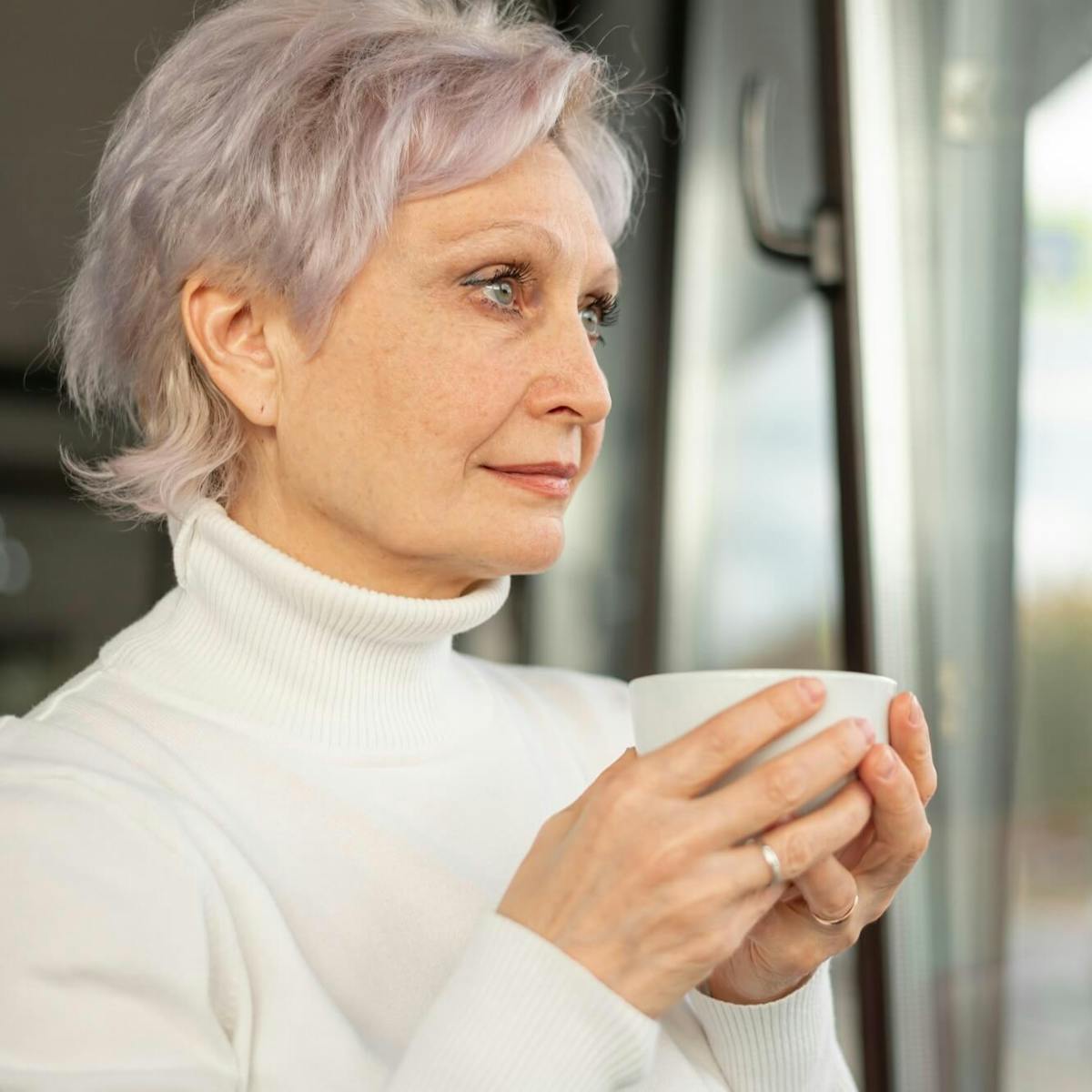 Ältere Frau steht mit einer Tasse in der Hand am Fenster und schaut heraus