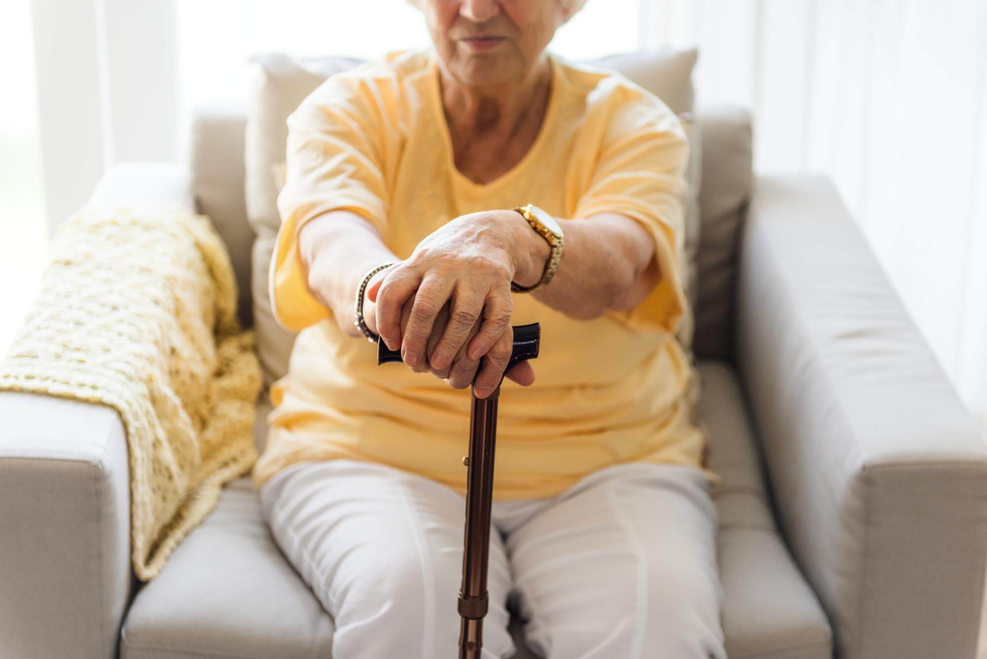 Ältere Frau mit Parkinson sitzt in einem Sessel und stützt ihre Hände auf einen Gehstock vor sich