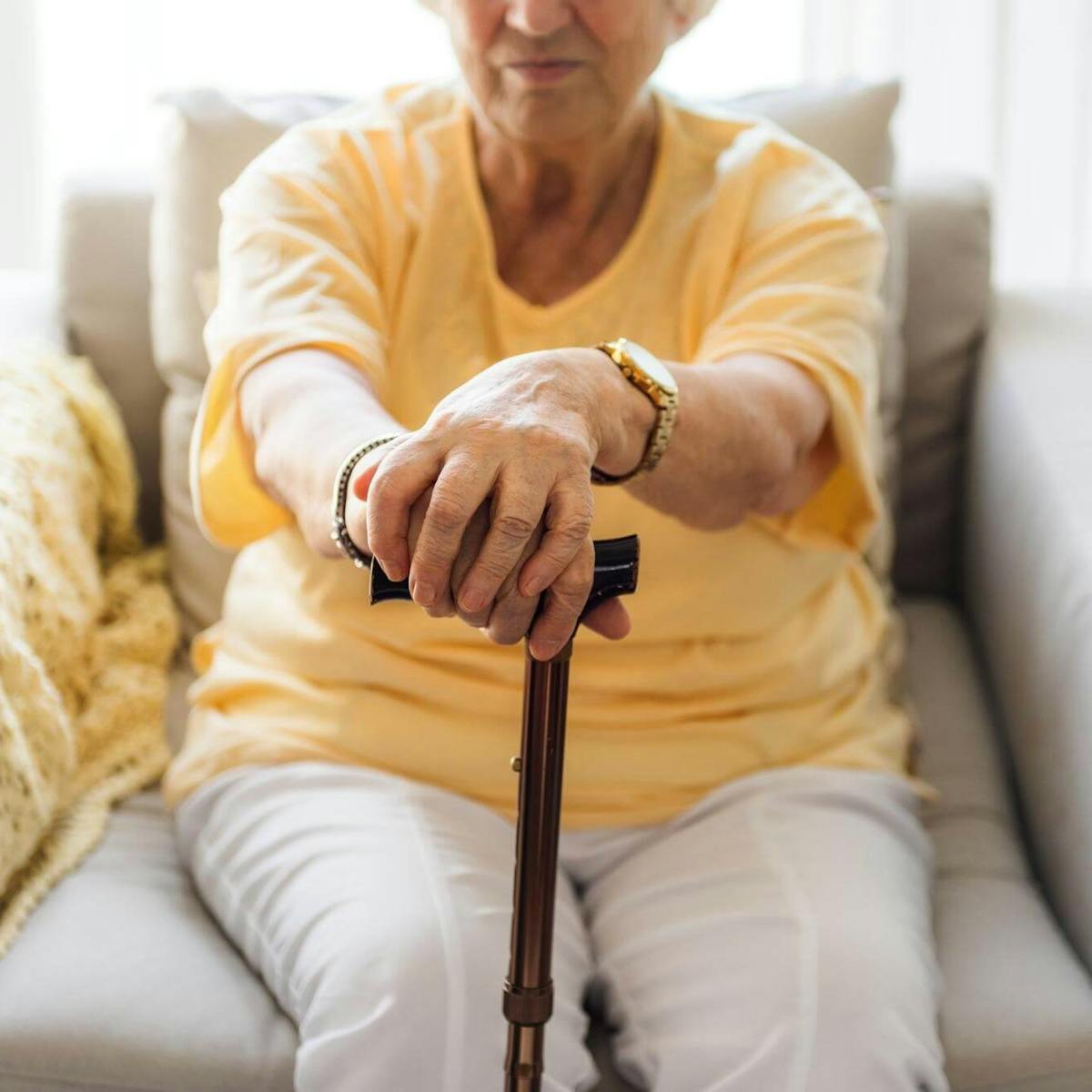 Ältere Frau mit Parkinson sitzt in einem Sessel und stützt ihre Hände auf einen Gehstock vor sich