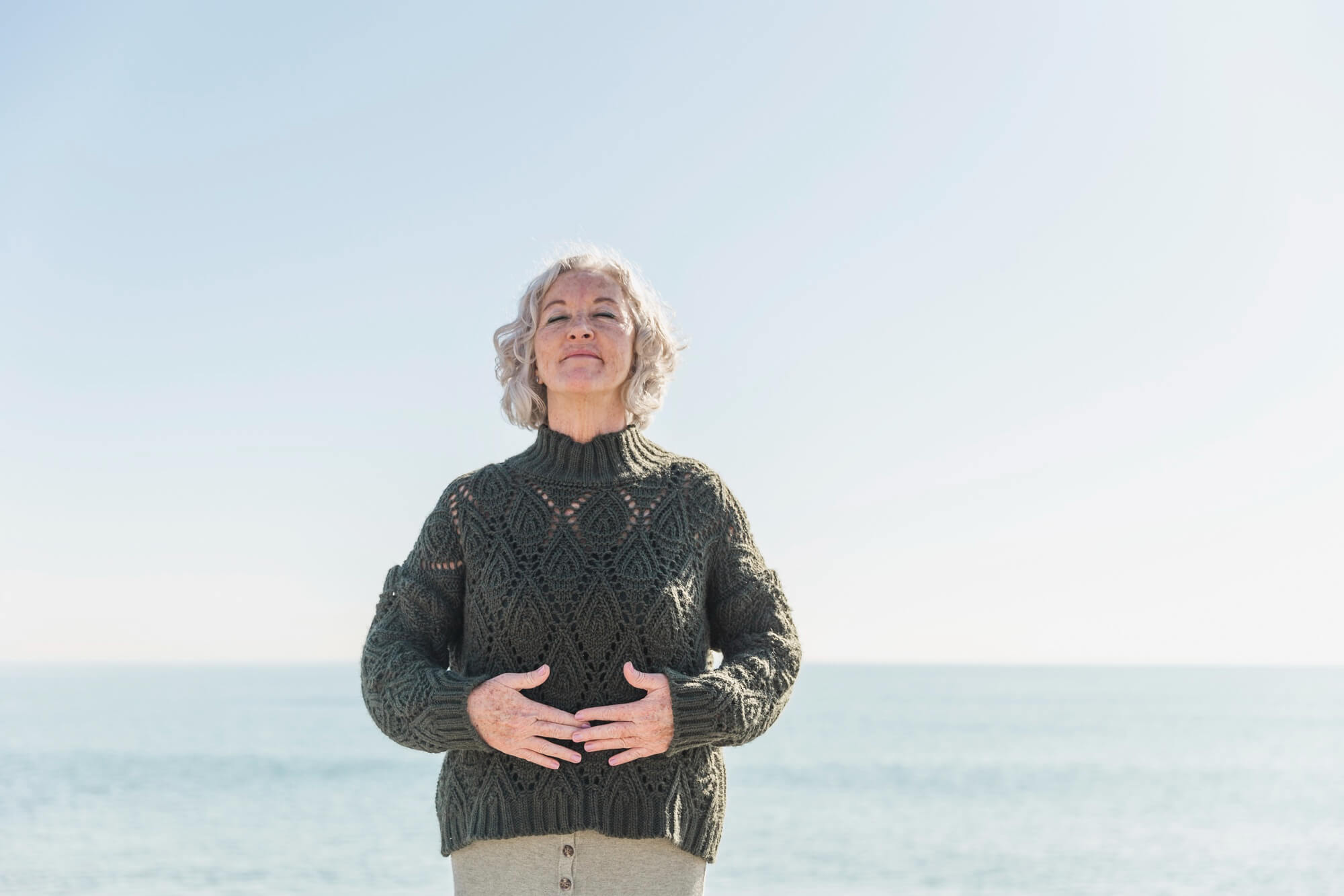 Frau steht vor dem Meer und hält sich die Hände vor den Bauch