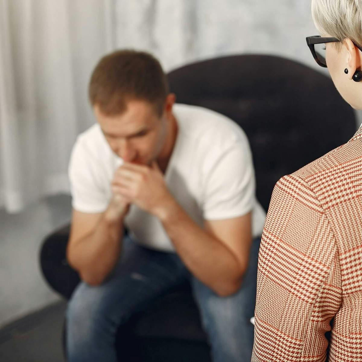 Frau steht vor einem Mann der ängstlich in einem Sessel sitzt und seinen Kopf auf die Hände stützt