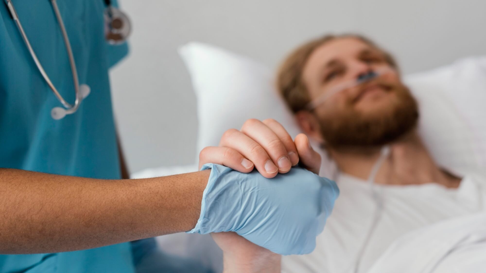 Männlicher Patient liegt im Krankenhausbett und hält die Hand einer Pflegeperson