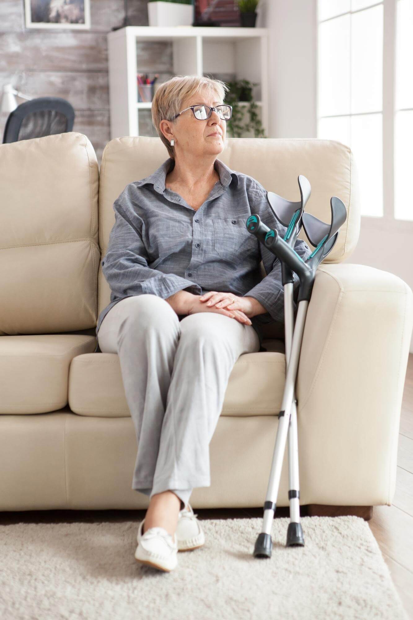 Ältere Frau sitzt auf dem Sofa, neben ihr sind Krücken an das Sofa gelehnt