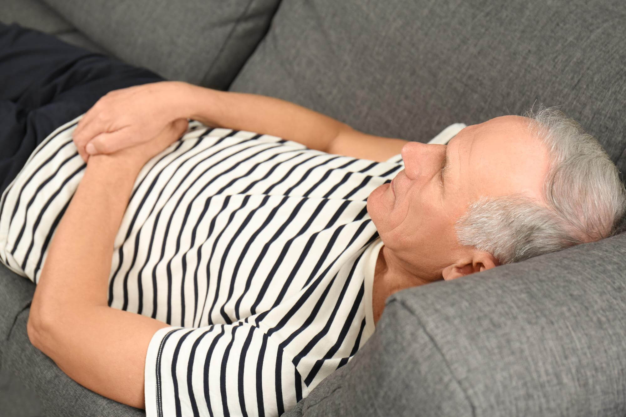 Älterer Mann im gestreiften Tshirt liegt auf dem Sofa und hält sich die Hände vor den Bauch
