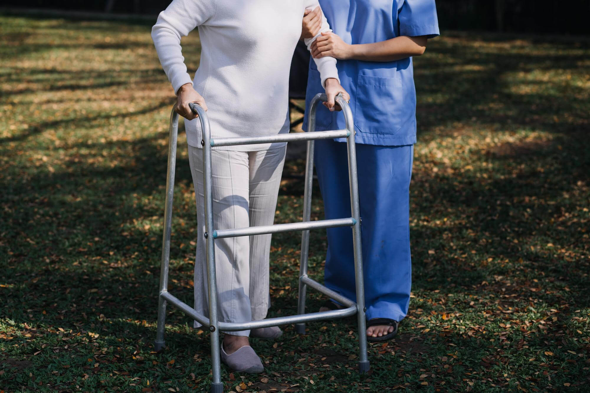 Osteoporose Patientin übt Gehen mit einer Gehhilfe auf einer Wiese während eine Pflegerin sie stützt