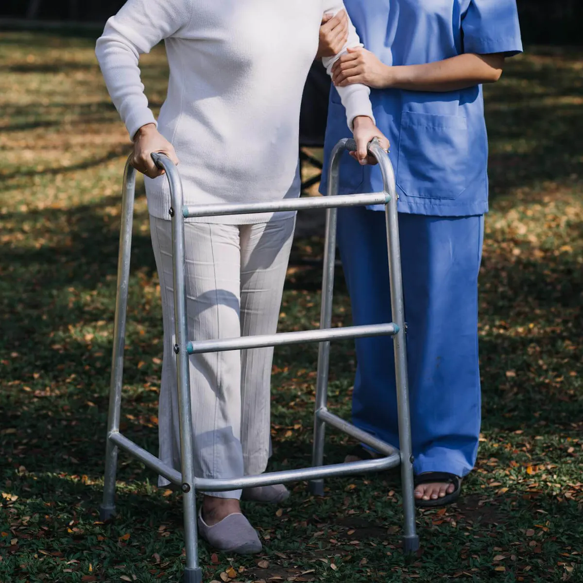 Osteoporose Patientin übt Gehen mit einer Gehhilfe auf einer Wiese während eine Pflegerin sie stützt