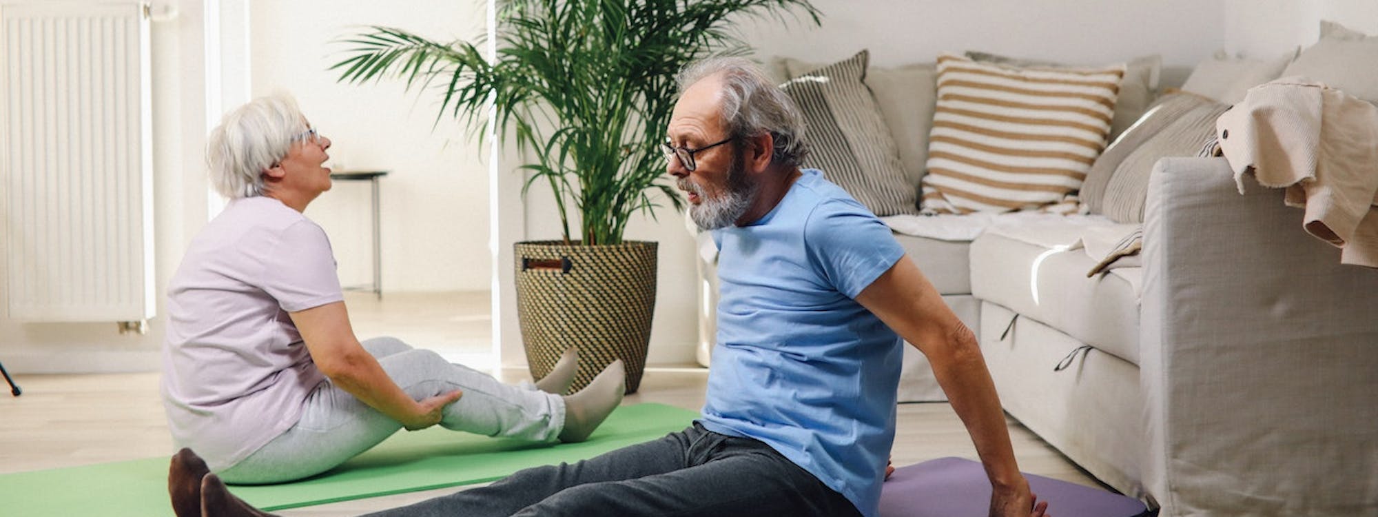 Eine ältere Frau und ein älterer Mann sitzen auf Sportmatten vor dem Sofa und dehnen sich