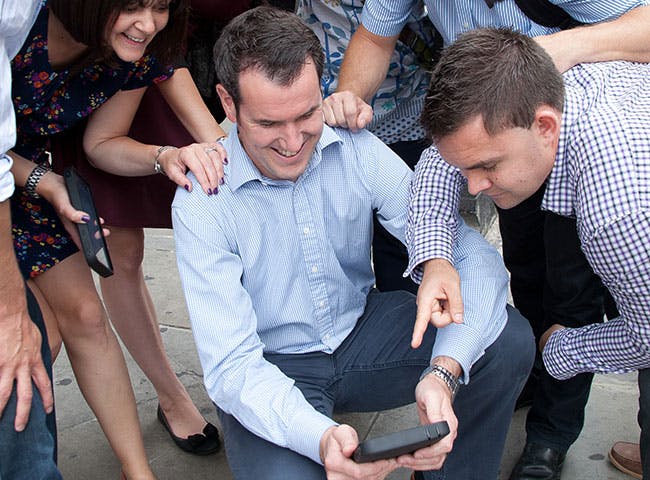 A man in office attire takes a selfie with his team gathered around him
