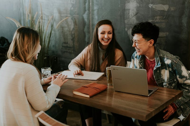 A group of coworkers smiling and laughing while looking at a laptop and notebook during a team meeting.