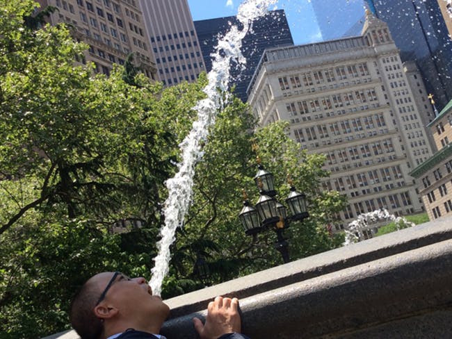 Perspective photo of a man pretending to drink water from a water fountain in the background