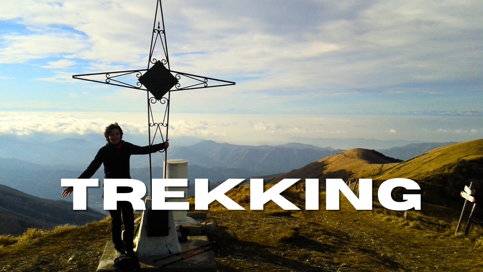 Persona accanto alla croce di vetta del Monte Ebro con nubi e crinali alle spalle