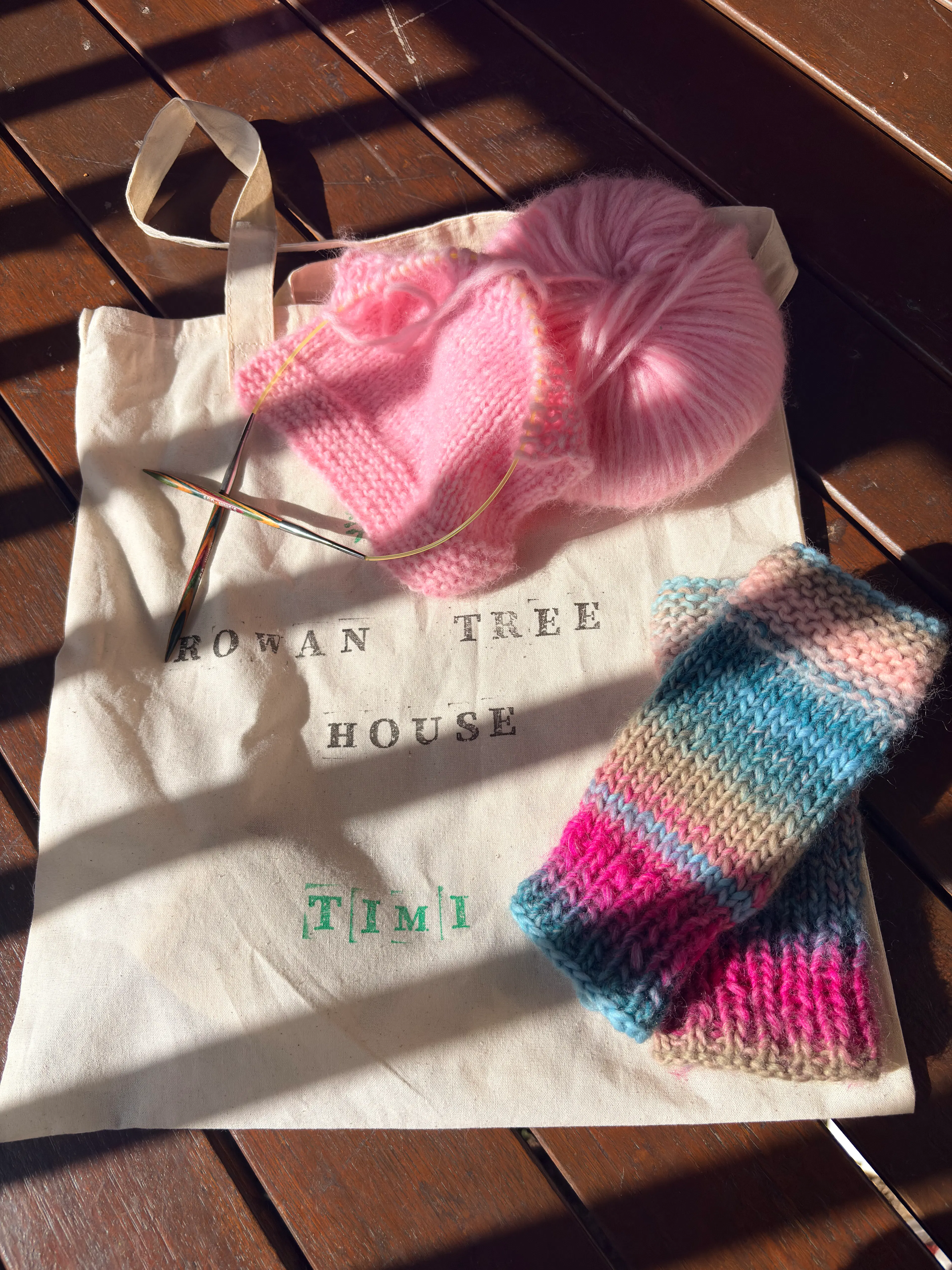 Knitted handwarmers on a personalised craft bag that says Rowan tree house, a knitting project in process, light pink yarn on bamboo rainbow knitting needles sits above. 